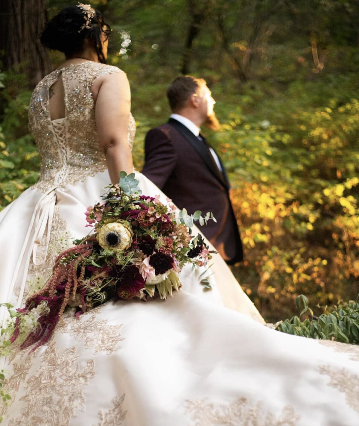 Bride and groom autumn portrait at Stone House Nevada City — bride in beaded gown with cascading dahlia and amaranth bouquet, groom in dark tuxedo, woodland setting
